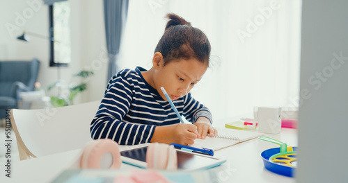 Asian toddler girl with sweater sit in front of desk with notepad use pencil focus on write notebook do homework from online learning course on the weekend at home. Distance online learning concept. photo