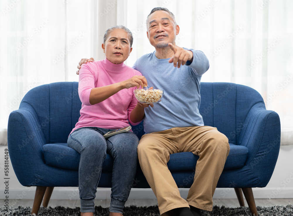 Senior couple sitting on sofa in living room have fun watching television with excited. Cheerful Asian elderly man and woman happy excitement watching tv entertainment. Family relaxation together. Senior couple sitting on sofa in living room have fun watching television with excited. Cheerful Asian elderly man and woman happy excitement watching tv entertainment. Family relaxation together.