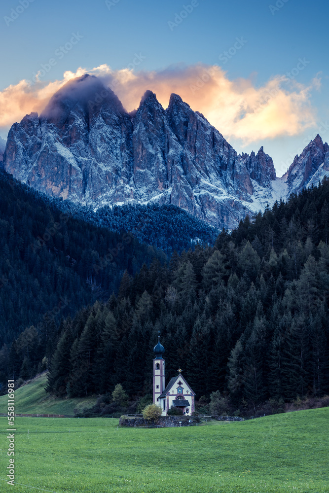 Farbige Wolken in Felsmassiv und kleine Kirche auf Wiese im Vordergrund in den Dolomiten.