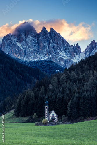 Farbige Wolken in Felsmassiv und kleine Kirche auf Wiese im Vordergrund in den Dolomiten.