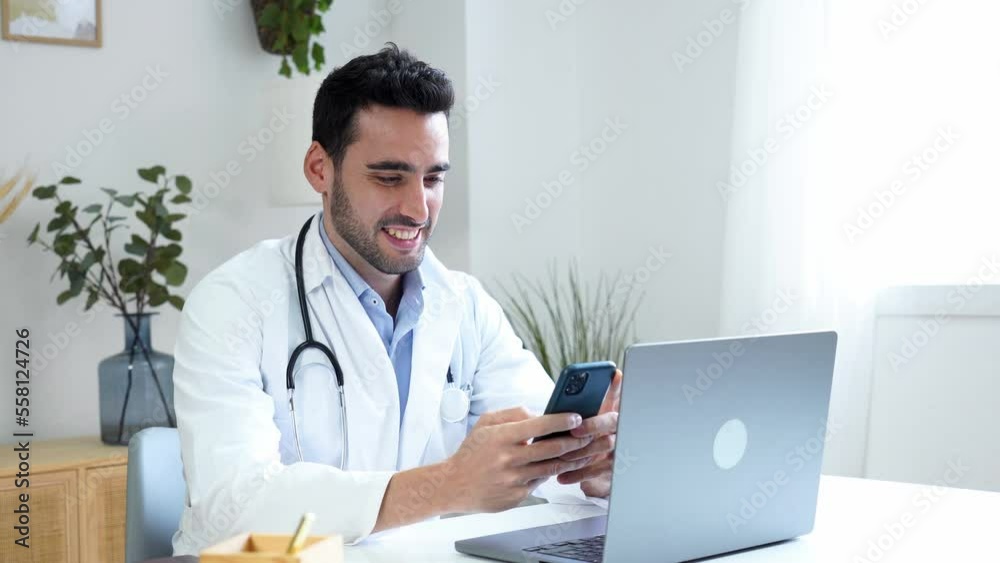 Positive Hispanic male doctor in medical uniform sitting at table with laptop and browsing mobile phone while working in modern clinic Positive Hispanic male doctor in medical uniform sitting at table with laptop and browsing mobile phone while working in modern clinic