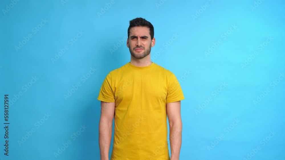 Young bearded male in yellow shirt looking at camera and doing a disappointment gesture while standing against blue background Young bearded male in yellow shirt looking at camera and doing a disappointment gesture while standing against blue background