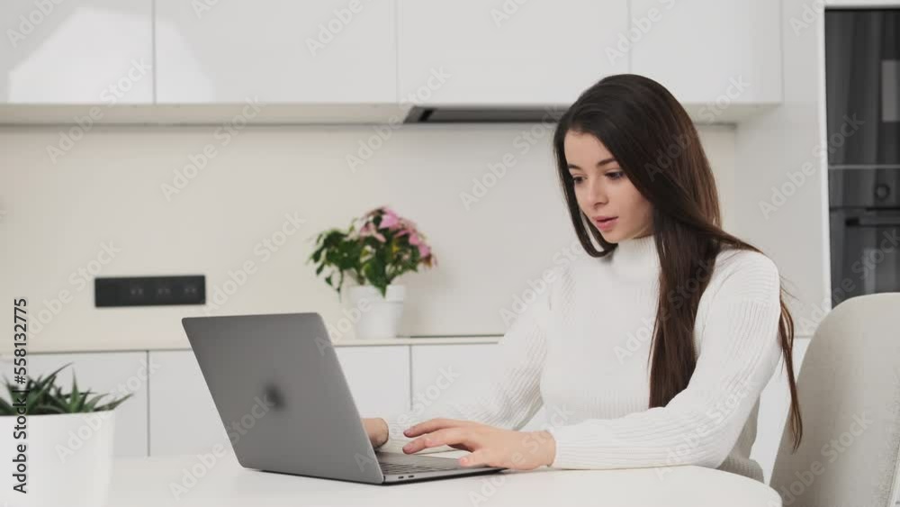 Brunette woman prepares report for university on Notebook sitting at desk in modern kitchen. Student gets surprised while typing on laptop Brunette woman prepares report for university on Notebook sitting at desk in modern kitchen. Student gets surprised while typing on laptop