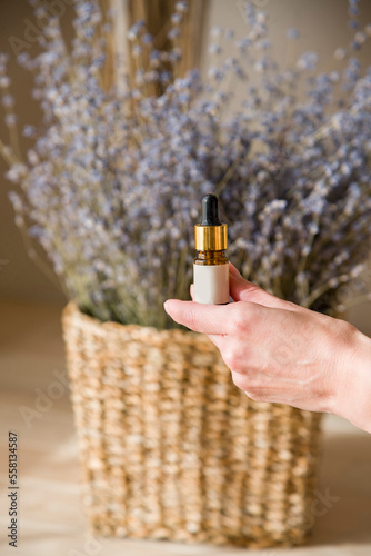 Woman's hands are holding a bottle with a pipette with lavender oil into the bottle on lavender background in straw basket
