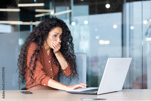 Fotografie Businesswoman has severe nausea, latin american woman at work sick, working sitting at table with laptop inside office