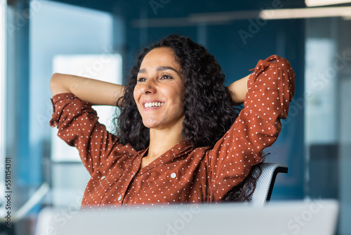 Happy and smiling successful hispanic business woman finished work satisfied with work result and achievement, hands behind head and smiling close up.