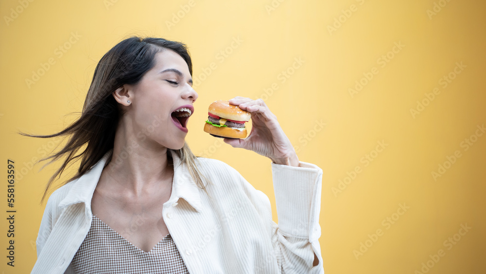 Cheat Meal Pretty Lady Eating Hamburger Junk Food. Cheerful Woman Smelling Burger Standing Over Yellow Background. Studio Shot Cheat Meal Pretty Lady Eating Hamburger Junk Food. Cheerful Woman Smelling Burger Standing Over Yellow Background. Studio Shot