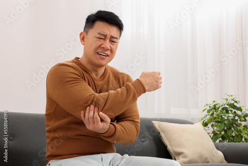 Asian man suffering from pain in his elbow on sofa indoors