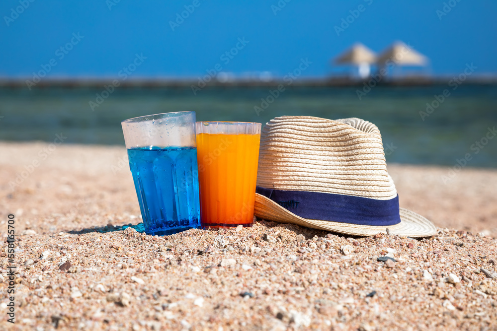 A hat and glasses with cocktails stand on the sand. Beautiful beach. Sunny summer day. Colorful cocktails in glasses. A hat and glasses with cocktails stand on the sand. Beautiful beach. Sunny summer day. Colorful cocktails in glasses.