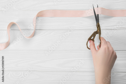 Woman cutting beige ribbon with scissors at white wooden table  closeup. Space for text