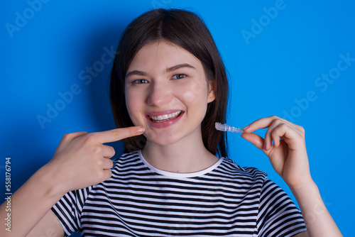 Young beautiful woman wearing striped t-shirt over blue background holding an invisible aligner braces and pointing at her perfect smile. 