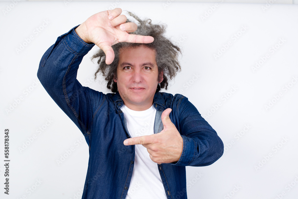Positive middle aged man with grey hair wearing denim jacket over white background with cheerful expression, has good mood, gestures finger frame actively at camera. Positive middle aged man with grey hair wearing denim jacket over white background with cheerful expression, has good mood, gestures finger frame actively at camera.