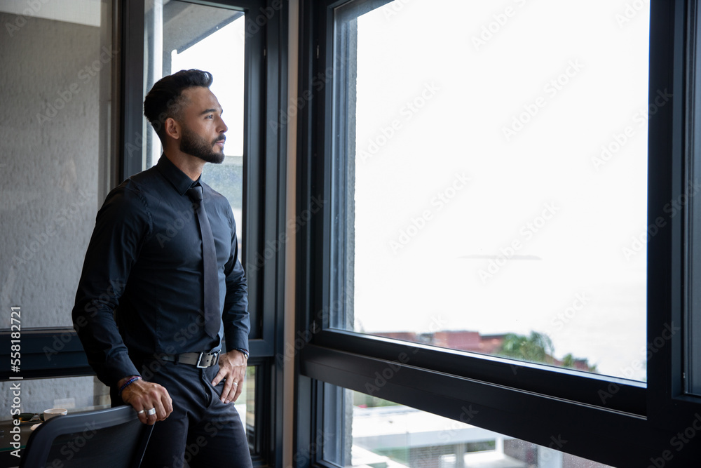 man standing by large window, young businessman standing in front of