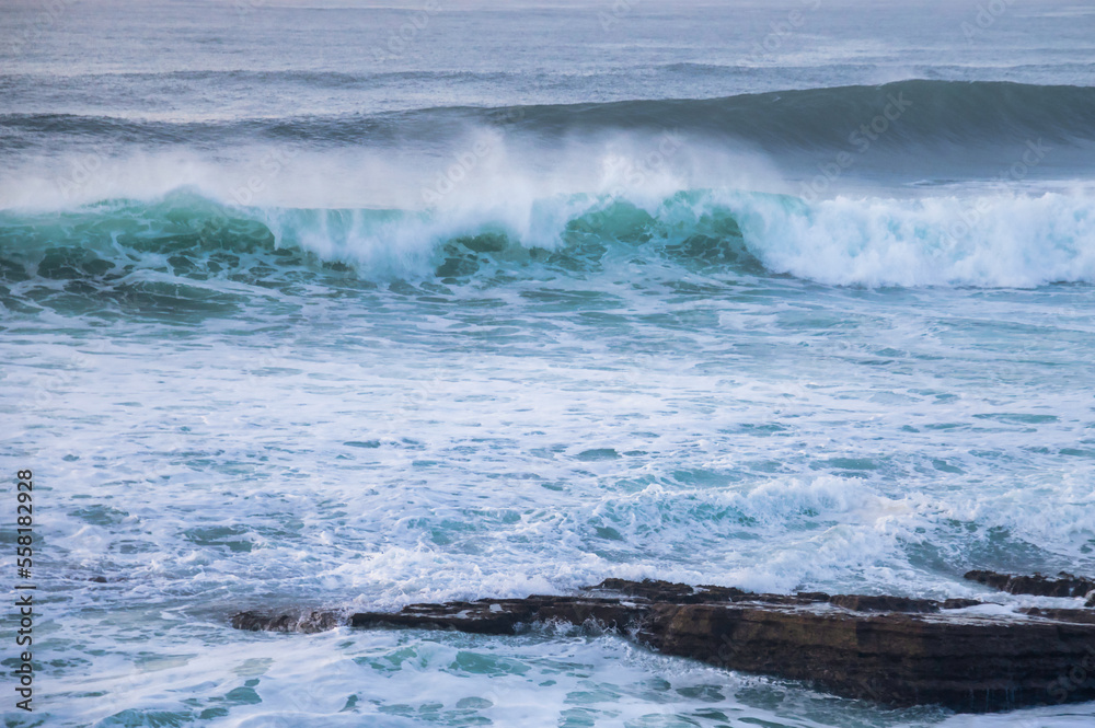 Fototapeta premium A powerful ocean wave crashes and breaks, creating foamy white spray as it approaches a rocky shoreline under an overcast sky. 