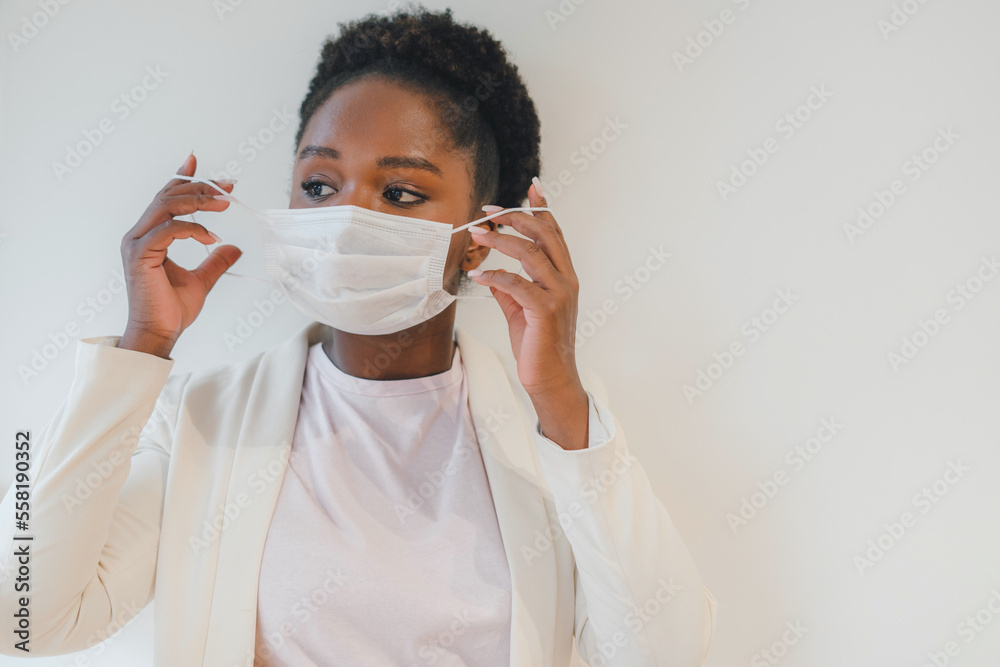 Portrait of african-american woman in white jacket putting on medical face mask and looking away isolated over with wall. Self isolation, prevention, quarantine Portrait of african-american woman in white jacket putting on medical face mask and looking away isolated over with wall. Self isolation, prevention, quarantine