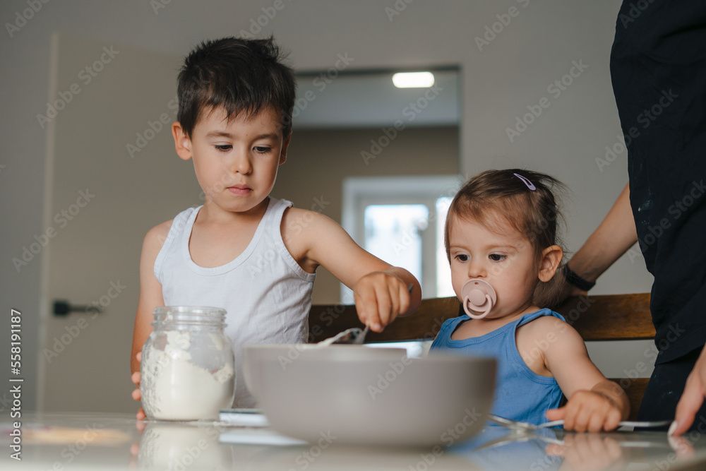 Two kids with their mother preparing dough for cookies together, adding flour and having fun in kitchen. Family education. Baking concept. Family relationships Two kids with their mother preparing dough for cookies together, adding flour and having fun in kitchen. Family education. Baking concept. Family relationships