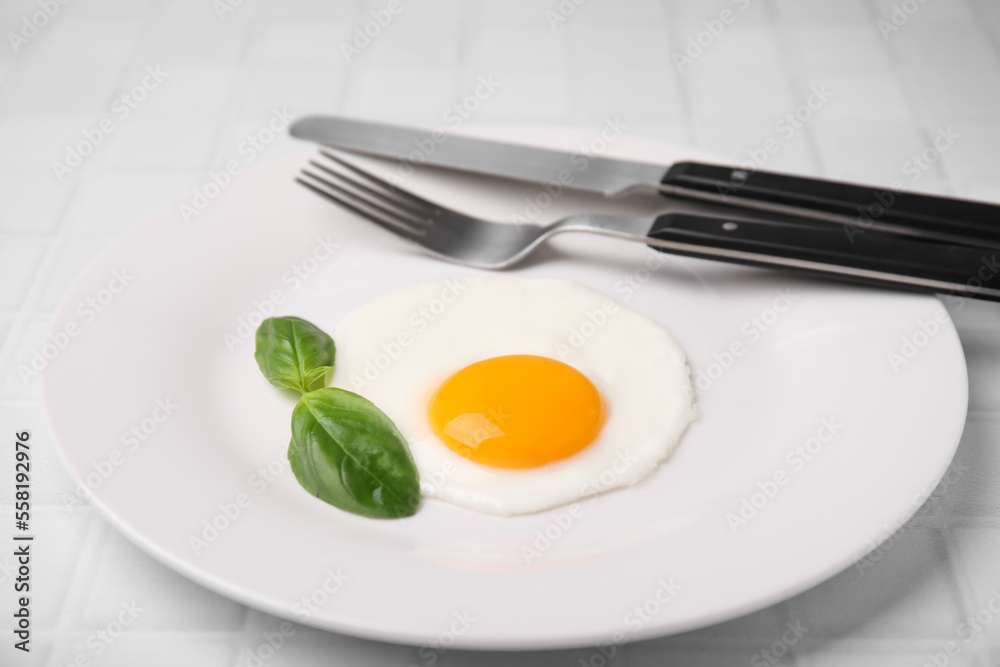 Tasty fried egg with basil in plate on white tiled table, closeup Tasty fried egg with basil in plate on white tiled table, closeup