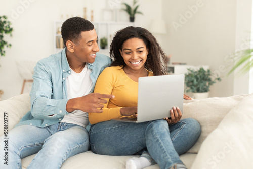 Glad millennial african american female showing laptop to male, recommend new app, video in living room
