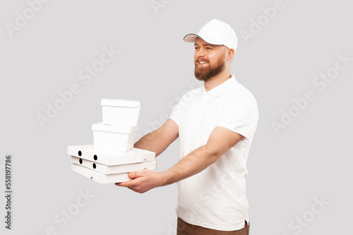 Image of young bearded delivery man in white uniform giving lunch boxes and pizza for custumer over grey background. photo