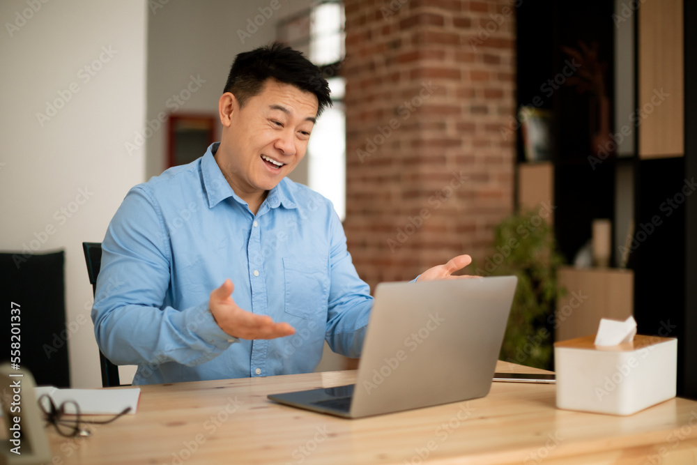 Excited asian mature man using modern laptop, video calling and gesturing at webcamera, sitting at workplace at home Excited asian mature man using modern laptop, video calling and gesturing at webcamera, sitting at workplace at home