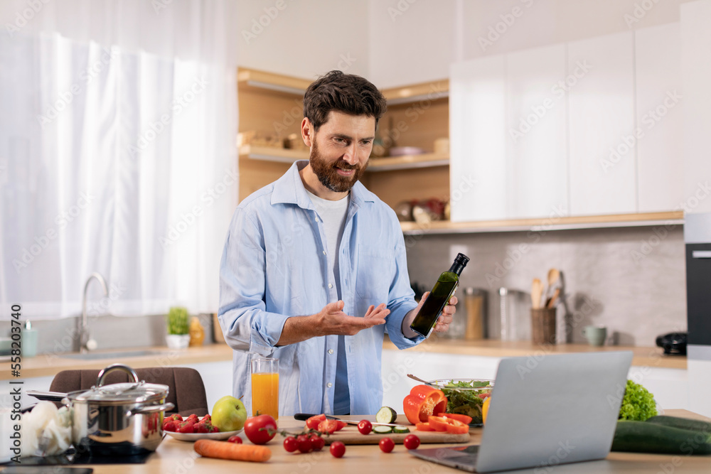 Smiling attractive millennial caucasian guy preparing eat at table with vegetables, show oil to computer webcam Smiling attractive millennial caucasian guy preparing eat at table with vegetables, show oil to computer webcam