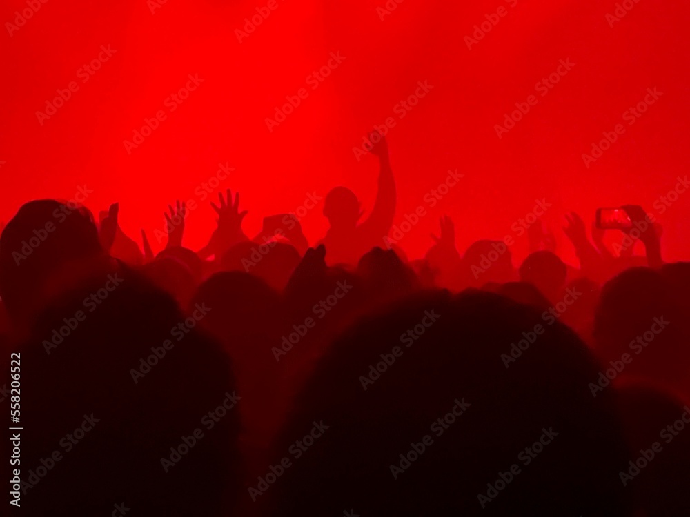 Rear view of large group of people enjoying a concert performance. There are many hands applauding. Red atmospheric spot lights firing from the stage. Rear view of large group of people enjoying a concert performance. There are many hands applauding. Red atmospheric spot lights firing from the stage.