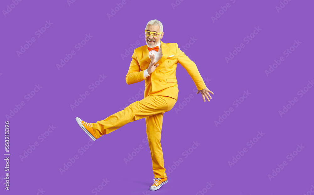 Overjoyed mature active man in yellow suit isolated on purple studio background dancing. Happy energetic old pensioner in jacket make dancer moves. Elderly party and celebration.