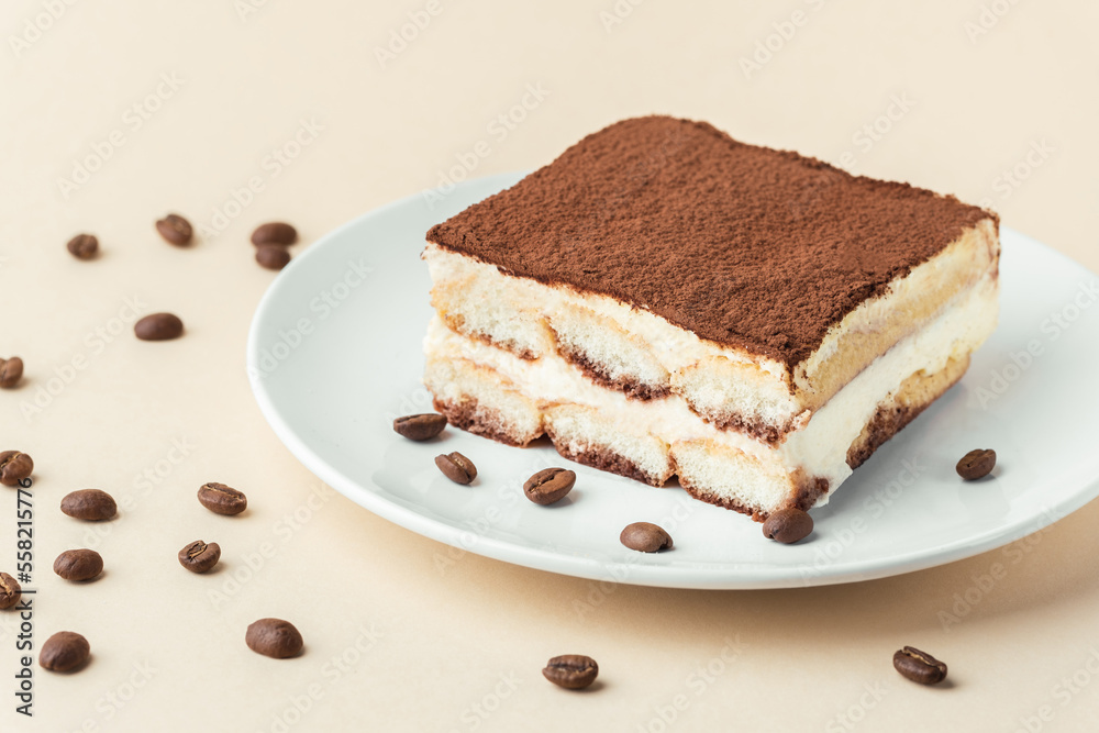 Homemade tiramisu cake on white plate against beige background surrounded by coffee beans . Italian dessert made of savoiardi cookies filled with mascarpone cheese and sprinkled with cocoa powder Homemade tiramisu cake on white plate against beige background surrounded by coffee beans . Italian dessert made of savoiardi cookies filled with mascarpone cheese and sprinkled with cocoa powder