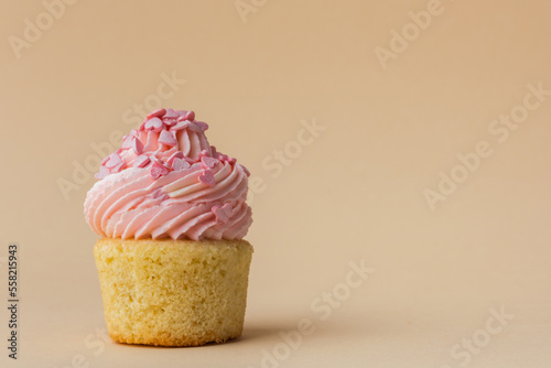 Close-up of vanilla cupcake with pink whipped butter cream top. Cream cheese frosting on muffin decorated with little pink heart shaped chocolate topping. Beige background. Happy Valentine's Day