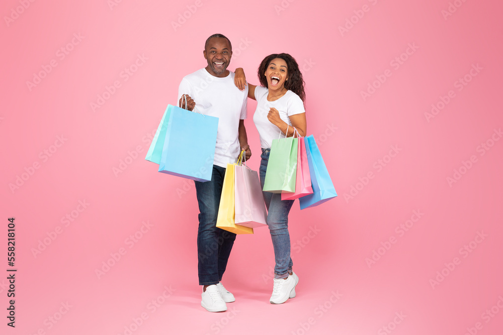 Happy african american man and woman holding colorful shopping bags and smiling at camera, posing over pink background