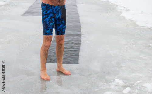 A seasoned strong man, his feet on the icy ice barefoot, dips into the hole, into the icy water in the lake, on a frosty winter day