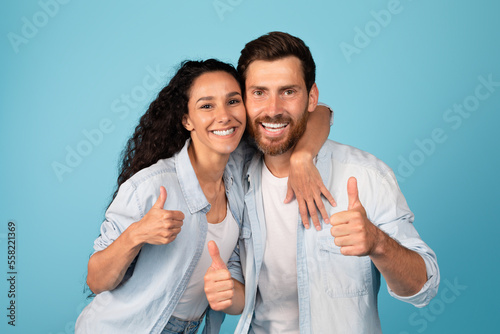 Cheerful young arab wife hugging european husband show thumbs up sign, have fun, enjoy success