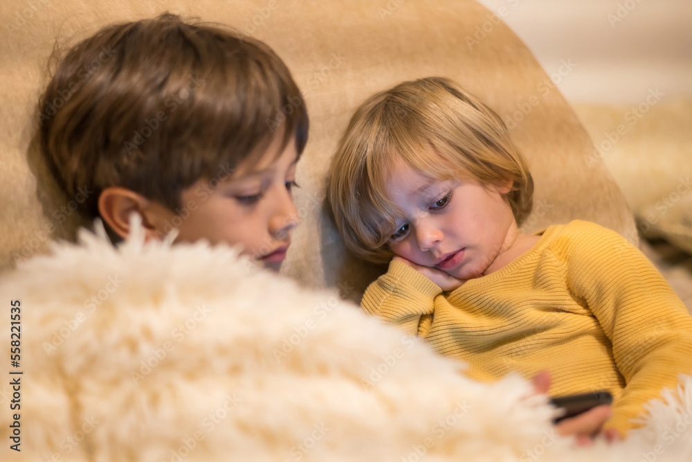 Two Beautiful Little Boys Sitting On The Sofa At Home. Siblings Entertained Watching And Playing With Smart Phone Inside The House.