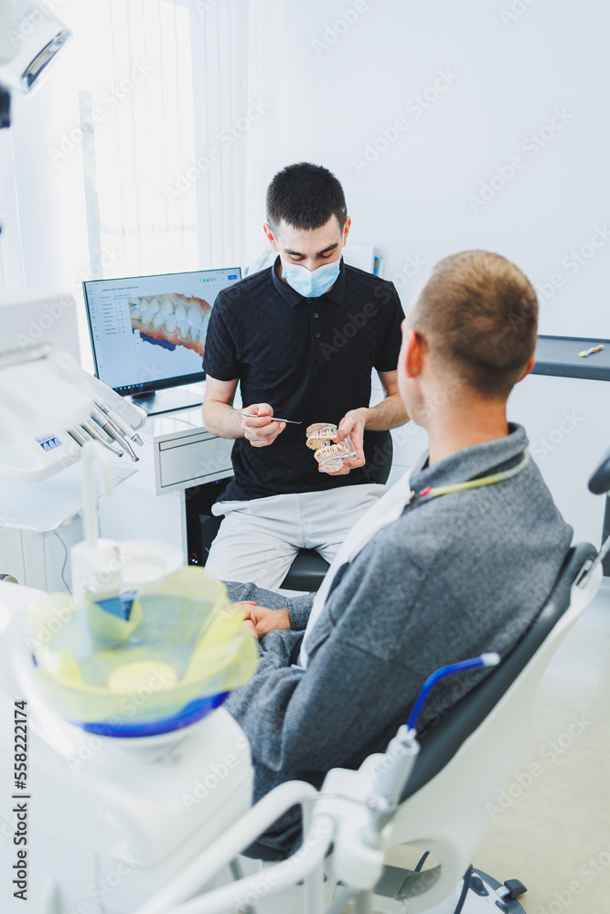 Obraz premium A dentist in a modern dental office tells a patient about dental care. A man sits in a dental chair and listens to a dentist.