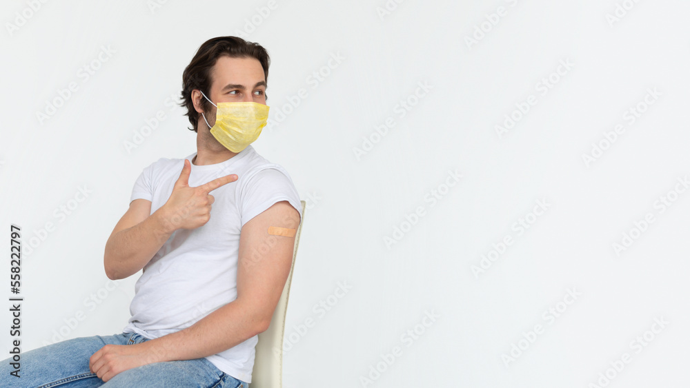 Glad young caucasian male in protective mask with band-aid on shoulder after vaccination shows finger on copy space Glad young caucasian male in protective mask with band-aid on shoulder after vaccination shows finger on copy space