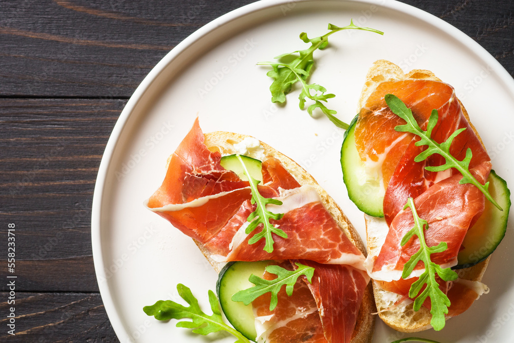 Open sandwiches with cream cheese, prosciutto, cucumber and arugula at wooden table.