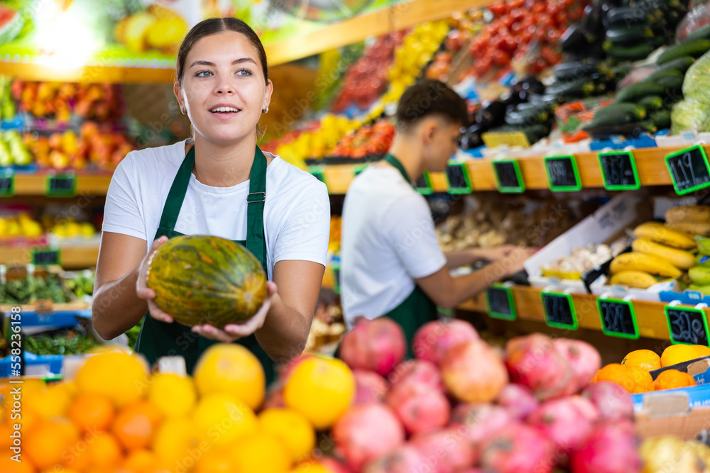 Positive young saleswoman holding fresh melon during offering fresh fruits in greengrocery store