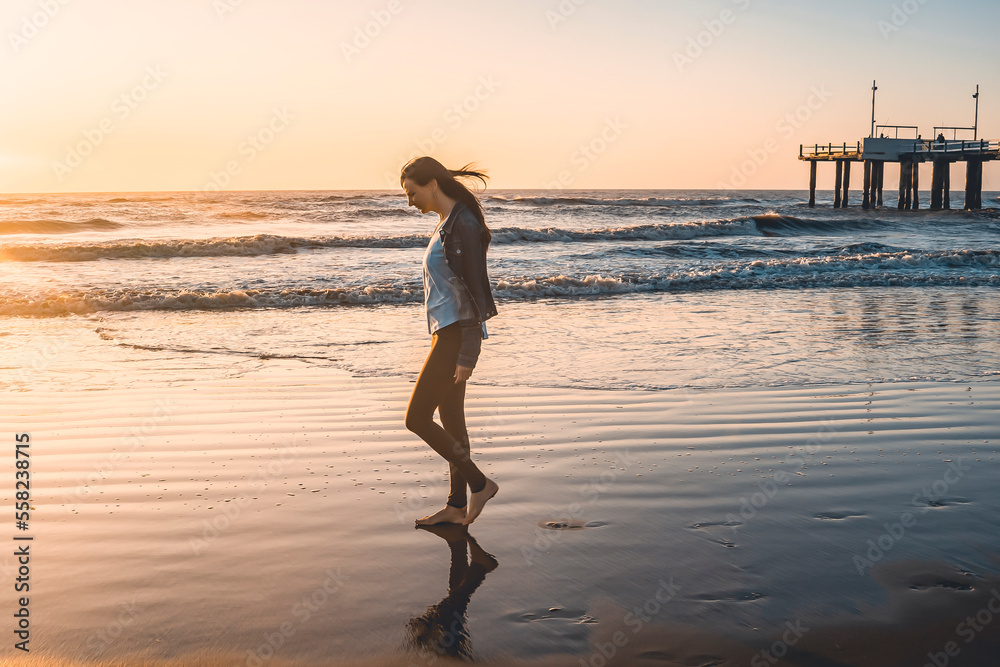 A lonely woman at the beach walking barefooted by the shore at sunrise.