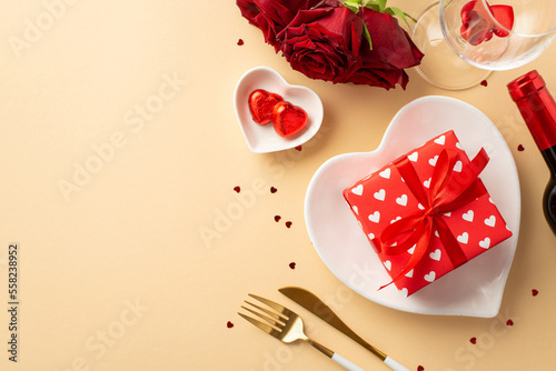 Valentine's Day concept. Top view photo of heart shaped dish with giftbox saucer with candies cutlery wine bottle glass red roses and confetti on isolated pastel beige background with empty space