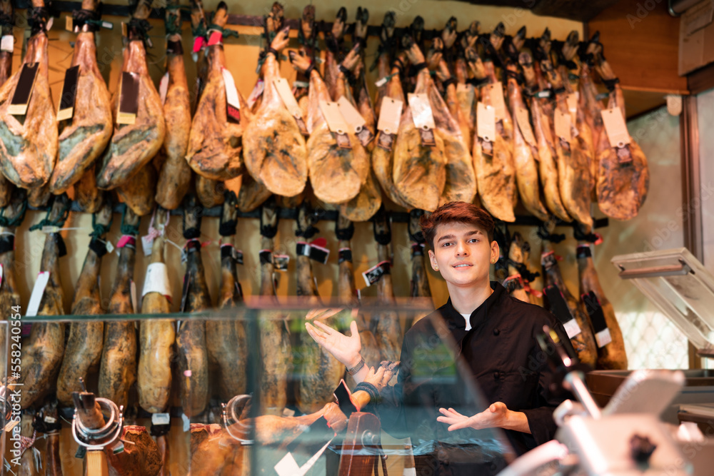 Portrait of smiling young salesman in black uniform working in local spanish meat shop, recommending delicious iberian ham