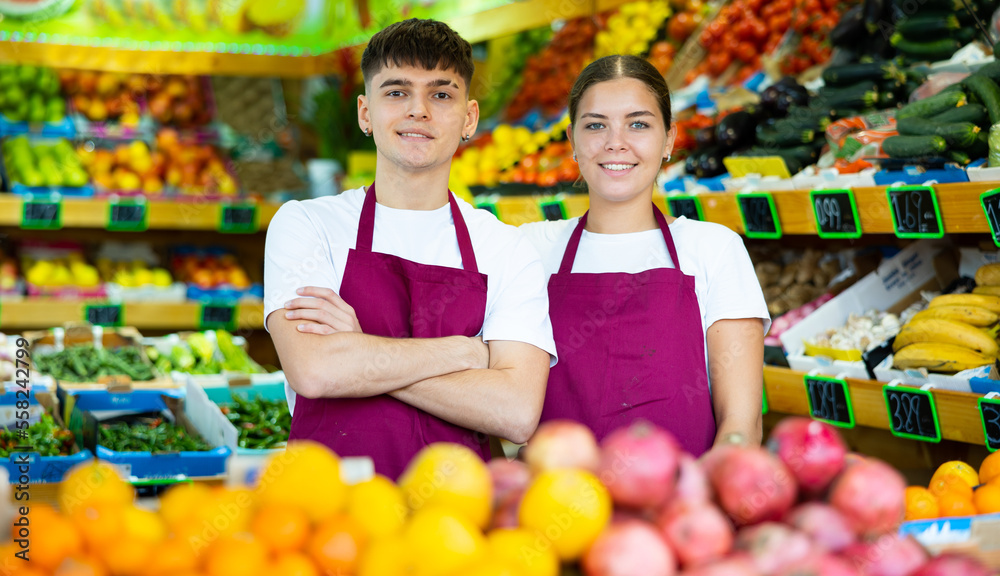 Portrait of young friendly confident male and female sellers wearing aprons and smiling at camera in fruit section of supermarket