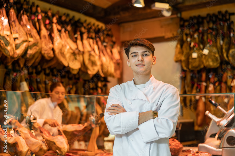 Portrait of experienced butcher on background of rack with hanging various jamon in shop