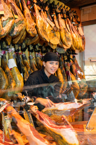 Attentive female seller cutting slices from whole leg of jamon in meat store