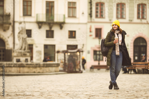 Coffee lover concept: full length portrait of smiling young woman drinking hot beverage at street of European city. Model wearing green coat, white, beige scarf, yellow beanie. Text space Outdoor shot