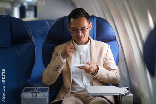 Satisfied millennial businessman drinking coffee, enjoying comfortable flight while sitting in airplane cabin.