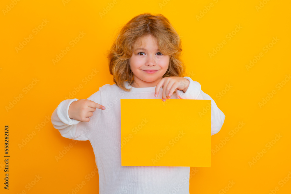 Cute child showing index finger on sheet of paper, isolated on yellow background. Portrait of a kid holding a blank placard, poster. Cute child showing index finger on sheet of paper, isolated on yellow background. Portrait of a kid holding a blank placard, poster.