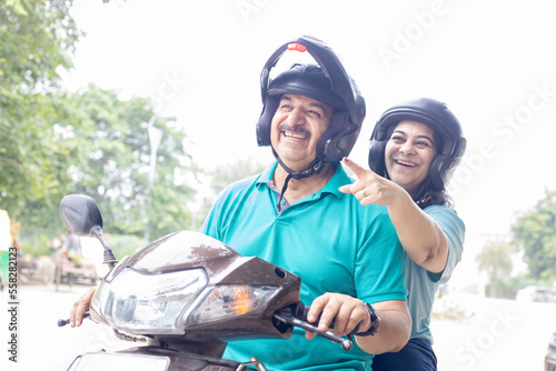 Happy senior indian couple wearing helmet riding motor scooter on road. Retirement life, Adventure and travel, Closeup © gajendra