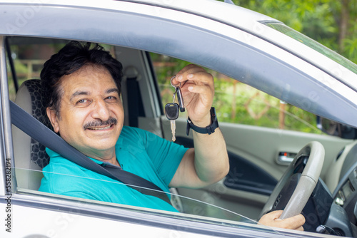 Happy indian mature man sitting in car wearing seat belt show key with hand ready to drive. Just buy new vehicle