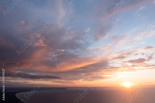 the sun is setting over the ocean with clouds in the sky and a plane flying in the sky over the water and land below it is a body of water with a boat in the foreground. .