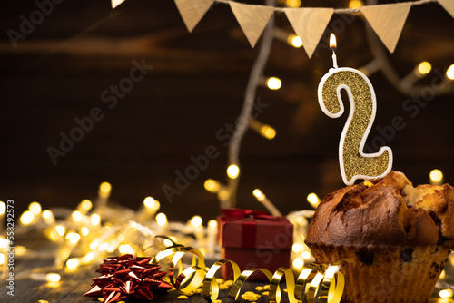 Number 2 gold burning candle in a cupcake against celebration wooden background with lights. Birthday cupcake. Copy space. photo